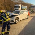 Feuerwehrleute reinigen die Fahrbahn nach einem Auffahrunfall. Im Hintergrund sieht man das havarierte Fahrzeug und ein Feuerwehrfahrzeug
